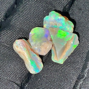 Könnte beinhalten: Vier rohe Opal-Edelsteine mit irisierenden Farbflecken in Grün, Blau und Gelb. Die Opale haben eine raue, natürliche Textur vor einem dunklen, strukturierten Hintergrund.