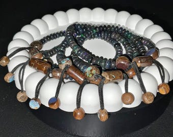 May include: A set of four black and iridescent opal beaded bracelets with brown wooden beads and black cords. The bracelets are displayed on a white circular stand.