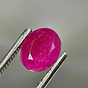 May include: A vibrant pink oval-shaped ruby gemstone held by tweezers.