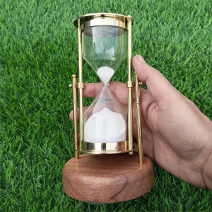 以下が含まれることがあります： 木製の台座と白い砂が入った真鍮製の砂時計。砂時計は緑の芝生の上に置かれています。