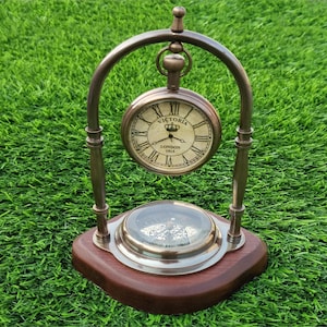 May include: A brass-toned metal desk clock and compass set mounted on a dark wooden base. The clock face features Roman numerals, the text VICTORIA, a crown icon, and the words LONDON 1915. A metal arch supports the clock above the compass.