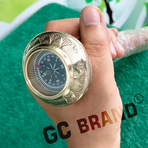 May include: A brass compass with an ornate design and a black face. The compass is mounted on a wooden walking stick. The compass face has markings for North, South, East, and West. The compass is labeled "GC BRAND".