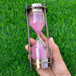 Op de afbeelding: Een zandloper van messing met een roze zandtimer. De zandloper is ongeveer 15 cm hoog en heeft een ronde basis.