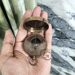 Puede incluir: Una brújula de latón con un reloj de sol y una cuerda atada a la parte superior. La brújula está abierta y el reloj de sol es visible. La brújula está en la palma de una mano.