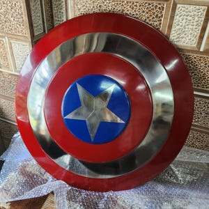 May include: A circular shield in red, silver, and blue, featuring a central star. The design includes concentric rings of red, silver, and blue. The centre has a blue circle with a silver star.