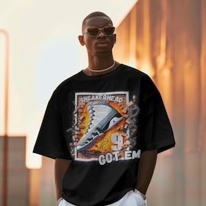 May include: A black t-shirt with a graphic of a white and grey sneaker breaking through a brick wall. The text "SNEAKERHEAD GOT 'EM" is printed on the shirt.