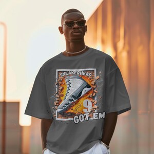 May include: A gray t-shirt with a graphic of a white sneaker breaking through a brick wall. The text "SNEAKERHEAD GOT 'EM" is printed on the shirt.