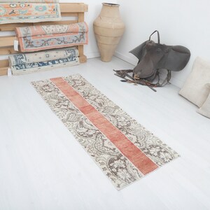 May include: A long, narrow runner rug with a white background and a paisley pattern in shades of brown. A wide, horizontal stripe of orange runs down the center of the rug.