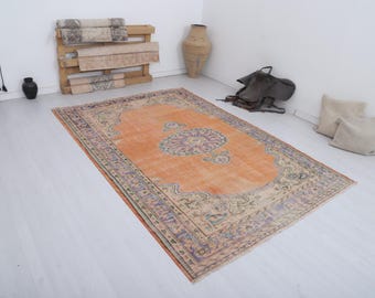 Puede incluir: Una alfombra vintage rectangular naranja y morada, con un medallón floral central y un borde detallado. La alfombra es de material tejido y está colocada sobre un suelo de madera clara. El diseño de la alfombra sugiere un estilo tradicional.