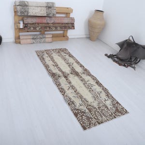May include: A long, rectangular rug with a cream central section and brown floral designs. The rug is laid on a light wooden floor. Other items in the room include a wooden pallet with rolled rugs, a vase, and a saddle.