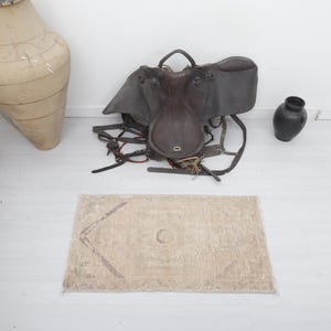 May include: A beige rug with a geometric pattern, a brown leather saddle, and a dark brown vase on a white floor.