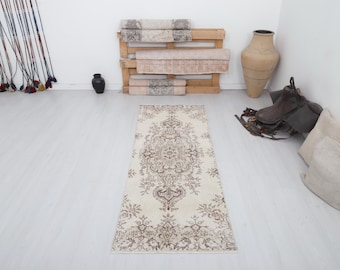 May include: A rectangular, cream-colored rug with an intricate, symmetrical brown floral pattern. The rug is placed on a light-colored floor, with other decorative items in the background.