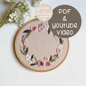 Puede incluir: Un bastidor de bordado floral con una corona de rosas rosas, flores blancas y hojas verdes. El bordado está sobre una tela beige, enmarcado por un aro de madera. El texto en la imagen dice "PDF & YouTube Video" y "revised & more detailed version".