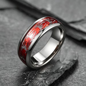 Può includere: Un anello in metallo color argento caratterizzato da una fascia lucida con un canale centrale incassato. Il canale contiene un inserto rosso vivace decorato con un motivo ripetitivo di sagome di ragni neri lungo tutta la circonferenza.