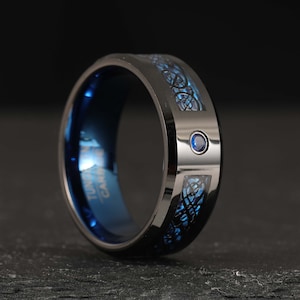 Op de afbeelding: Zwarte ring van wolframcarbide met een blauw Keltisch knoopontwerp en een kleine blauwe saffier.