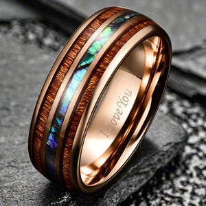 May include: A rose gold-toned ring featuring a central inlay of iridescent abalone shell flanked by two bands of dark brown wood grain. The interior of the polished metal band is engraved with the cursive text I Love You in a dark font.
