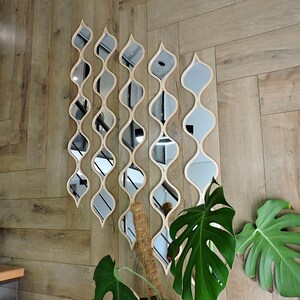 May include: A set of decorative wall mirrors in a unique, flowing design. Each mirror has a light wood frame and a series of curved, reflective surfaces. The mirrors are arranged in a vertical pattern on a wooden wall, with green plants in the foreground.