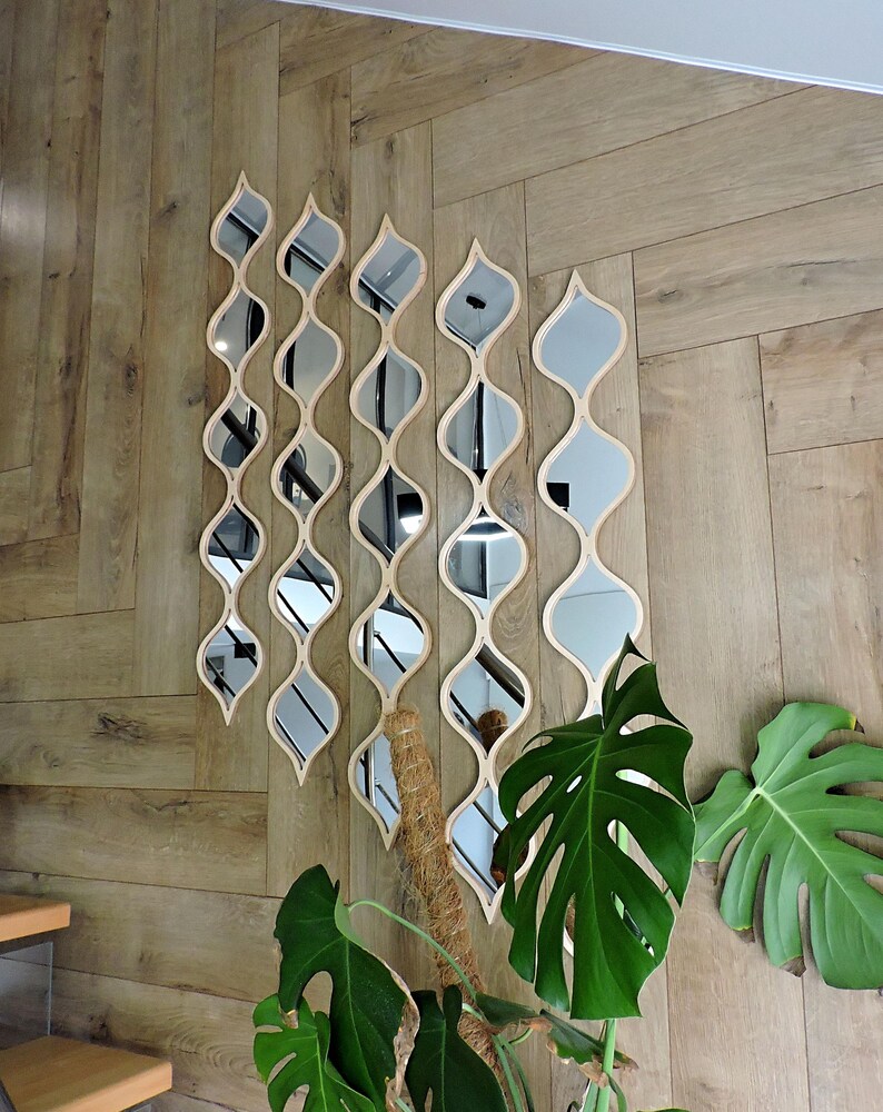 May include: A set of decorative wall mirrors in a unique, flowing design. Each mirror has a light wood frame and a series of curved, reflective surfaces. The mirrors are arranged in a vertical pattern on a wooden wall, with green plants in the foreground.