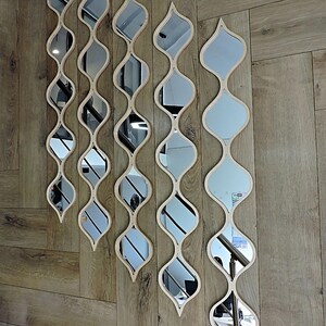 May include: A collection of decorative wall mirrors with a unique, flowing design. Each mirror features a series of interconnected, teardrop-shaped reflective surfaces, framed by light-colored wood. The mirrors are arranged vertically on a wooden wall, creating a stylish, modern aesthetic.