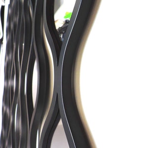 K&ouml;nnte beinhalten: Schwarze Wanddekoration aus Metall mit einem wellenf&ouml;rmigen, abstrakten Design. Die Dekoration hat eine verspiegelte Oberfl&auml;che in der Mitte.