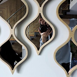May include: A set of decorative wall mirrors with a unique, flowing design. Each mirror is framed with light-colored wood in a repeating, stylized teardrop shape. The mirrors reflect the surrounding room, including a staircase and a person holding a camera.