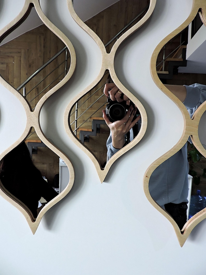 May include: A set of decorative wall mirrors with a unique, flowing design. Each mirror is framed with light-colored wood in a repeating, stylized teardrop shape. The mirrors reflect the surrounding room, including a staircase and a person holding a camera.