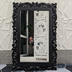 May include: A rectangular black mirror with an elaborate, carved frame. The frame has detailed floral and scrollwork patterns. The mirror reflects a room with a white screen with floral cutouts and a person holding a phone.