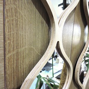 May include: A decorative wall mirror with a unique design. The mirror features a light wood frame with a layered, wavy pattern. The background is a wooden wall with vertical grain, and the mirror reflects the surrounding environment.
