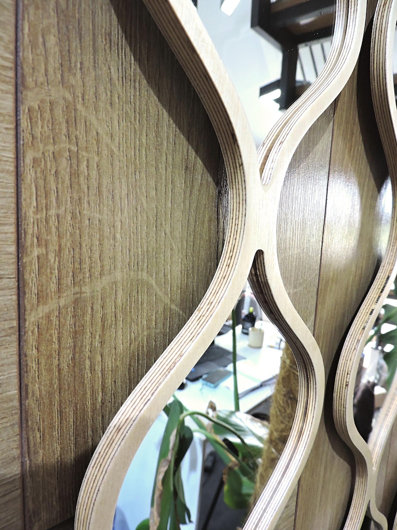 May include: A decorative wall mirror with a unique design. The mirror features a light wood frame with a layered, wavy pattern. The background is a wooden wall with vertical grain, and the mirror reflects the surrounding environment.