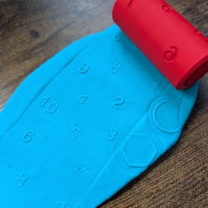 May include: A bright red cylindrical rolling pin with embossed numbers and shapes is pressed into a flat sheet of light blue modelling clay. The clay shows clear, indented impressions of various numbers and geometric shapes left by the roller.