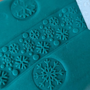 May include: Teal blue play dough with snowflake designs pressed into it. The snowflake designs are created using a white plastic roller with snowflake patterns.