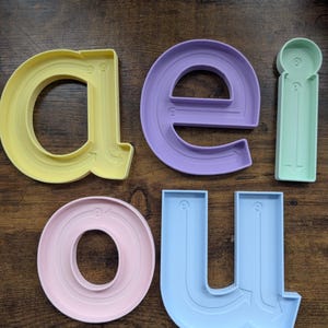 May include: A set of five plastic lowercase vowel letter forms in pastel colours. The yellow 'a', purple 'e', light green 'i', pink 'o', and light blue 'u' each feature embossed directional arrows and numbered guides to demonstrate the stroke order for writing.