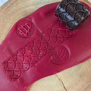 May include: A red clay surface with a pattern of scales, a Christmas tree, and a snowflake. A brown 3D printed roller with a scale pattern is on top of the clay.