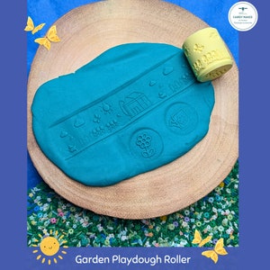 May include: A teal playdough with garden-themed imprints, including a house, sun, and flowers, is on a wooden slice. A yellow roller with garden designs is next to the playdough. The words "Garden Playdough Roller" are at the bottom.