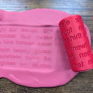 May include: A cylindrical red rolling pin with embossed text featuring number words like six, eleven, one, four, twelve, nineteen, thirteen, seven, two, twenty, sixteen, fifteen, seventeen, three, five, ten, and fourteen pressed into pink clay.