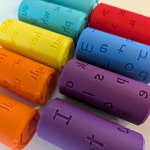 May include: A set of colourful, cylindrical blocks with letters and numbers printed on them. The blocks are arranged in a rainbow pattern, with red, orange, yellow, green, blue, and purple blocks.