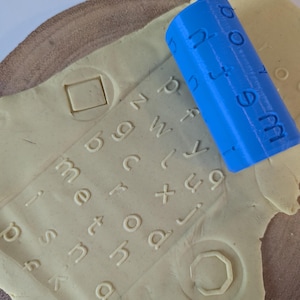 May include: A blue cylindrical stamp tool with letters and numbers is pressed into a light-coloured clay surface. The word "method" is visible. The clay also has a square and a hexagon shape pressed into it.