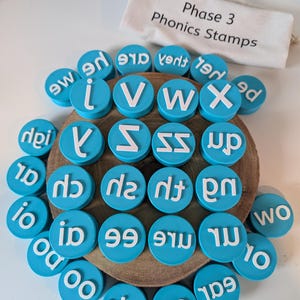 May include: A set of blue, circular stamps with white lettering, displayed on a wooden disc. The stamps feature letters and words such as "i", "V", "W", "X", "up", and "wo". A small canvas bag with the text "Phase 3 Phonics Stamps" is visible in the background.