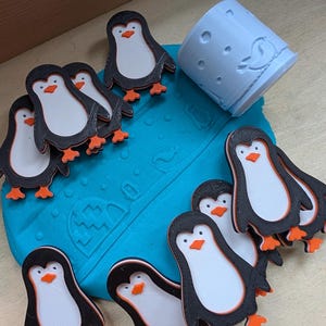 May include: A group of penguin cutouts with black bodies, white bellies, and orange feet and beaks. A light blue cylindrical object with a penguin design is also present. The penguins are arranged on a blue surface.