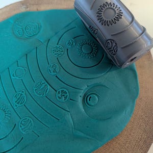 May include: A grey roller with a solar system design is pressed into teal playdough. The playdough shows an imprint of the sun and planets. The roller has a cylindrical shape with detailed engravings of celestial bodies.