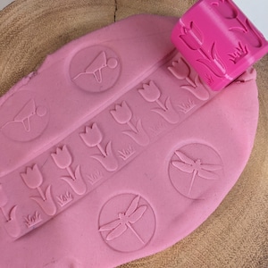 May include: Pink play dough with various impressions made by a pink rolling pin with a tulip design. The impressions include tulips, dragonflies, and wheelbarrows.