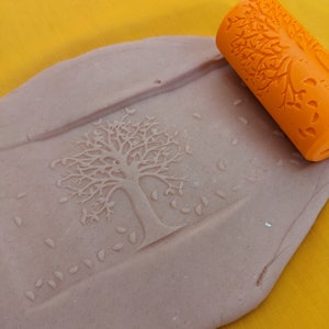 May include: Orange rolling pin with a tree design used to create an impression of a tree with leaves in a light brown clay.