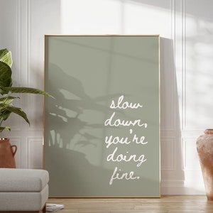 Puede incluir: Una impresión enmarcada de color verde salvia con el texto "slow down, you're doing fine." La impresión está colgada en una pared blanca con una planta en maceta y dos macetas de terracota en primer plano.