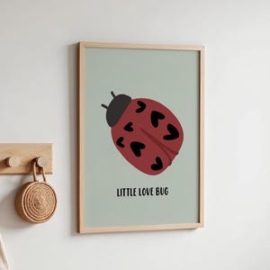 Może przedstawiać: Oprawiony wydruk przedstawiający czerwoną biedronkę z plamkami w kształcie serca i napisem "LITTLE LOVE BUG" na jasnym zielonym tle. Scena przedstawia pokój dziecięcy z drewnianym wieszakiem, ubrankami dziecięcymi i łóżeczkiem.