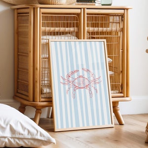 May include: A framed print with a blue and white striped background and a red crab illustration. The print is leaning against a light wood cabinet with a woven rattan design.