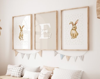 May include: Three framed wall art prints with a bunny theme. The prints feature watercolor illustrations of bunnies and a monogram. The frames are light wood, and the overall color scheme is neutral with beige and white tones. A woven giraffe and storage bench are also in the room.