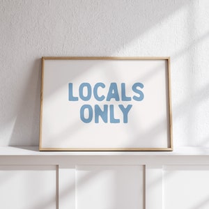 Op de afbeelding: Een ingelijst afdruk met de tekst "LOCALS ONLY" in blauwe letters op een witte achtergrond.