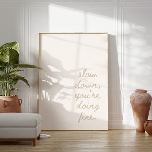 May include: A beige poster with the text "slow down, you're doing fine." hanging on a white wall with a potted plant and two terracotta pots in the foreground.