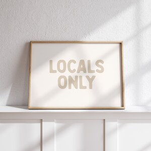 May include: A framed print with the words "LOCALS ONLY" in a beige font. The print has a light beige background and a natural wood frame. The print is displayed on a white shelf against a white wall.