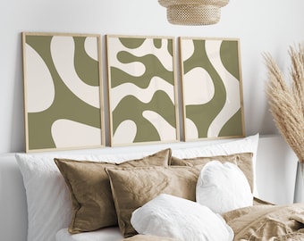 May include: Set of three framed abstract art prints. Each print features a unique design in olive green and off-white, with a natural wood frame. The artwork is displayed above a bed with neutral-toned bedding.